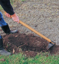 Person in calf-length rubber boots - jeans & dark sweater using long-handle trench digger in brown soil. Trench digger looks like a hoe but with more of a cupped blade