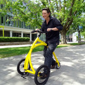 Person dressed in black on a yellow Alinker Walking Bike on a tree-shaded sidewalk