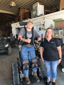 Kane in stand-up tracked wheelchair giving thumbs up and standing next to Rachel Jarmon in farm shop.