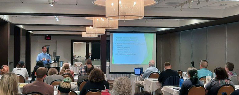 Male presenter at podium in hotel meeting room with presentation on screen to right and approximately 20 seated participants in foreground