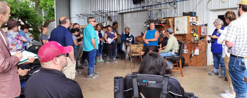 Male staff member talks with male farmer, both seated, in large farm shop with approximately 20 people gathered around listening