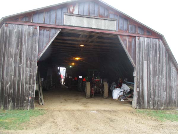 Pictured is a large, old wooden machinery/materials-storage building, its double sliding doors pulled open.