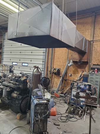 Pictured inside this equipment storage/maintenance building and directly above a metalworking table is a ceiling-suspended, 3-foot-square ventilation hood that exhausts to the outside.