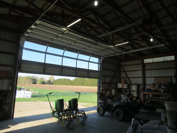 Pictured (from inside looking out) is a farm building’s wide, segmented, glass-paneled overhead door that’s partially raised, a small towed seeder at the just-opened entrance.