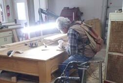 Pictured is an adult male seated at his workbench, a 3-foot-long adjustable-height LED lamp providing excellent lighting for the meticulous task he’s carrying out.