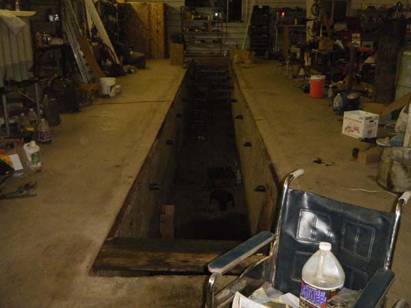 Pictured is a workshop vehicle/machinery service pit (about 4-feet wide/20 feet long) with access steps at both ends and metal crossbar anchors near the top of both sides about 3 feet apart.