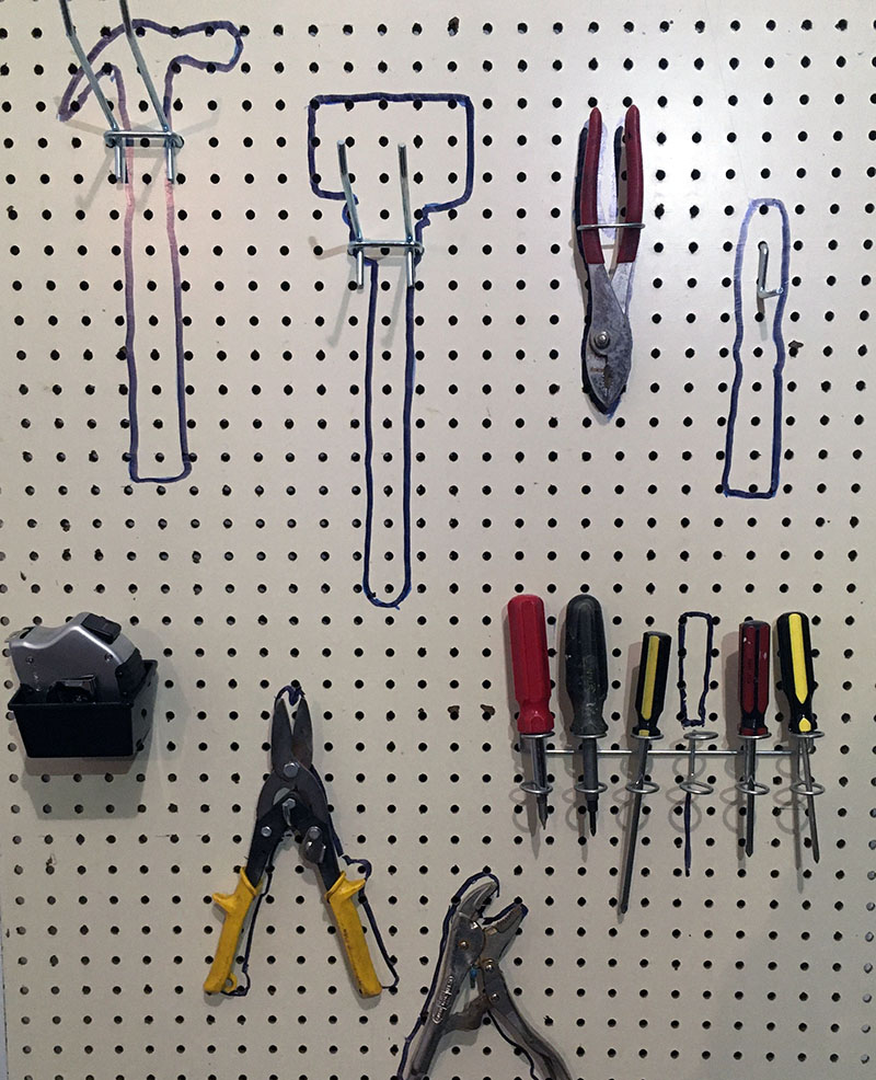 Pictured is a portion of wall-mounted peg board, its hooks holding hammer, mallet, and screw drivers. etc.; drawn behind each is an outline of that particular tool.