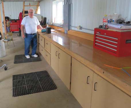 Pictured is an adult male standing on one of two 2-x 4-foot cushioned floor mats in front of his workbench. 