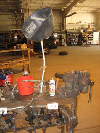 Pictured is a welding table with two electrical outlets on one of its long sides, a vise at one end, tools underneath, various items on top, and a face shield suspended just above the table.