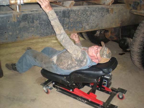 Pictured is an adult male reclined on a creeper (its battery-powered cushioned backrest in partially raised position) performing maintenance under a grain wagon.