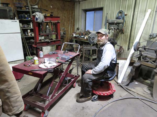 Pictured next to the 4-wheeled, 5-foot-long, hydraulic scissors-lift cart is an adult male seated on his adjustable-height, wheeled work stool with backrest.