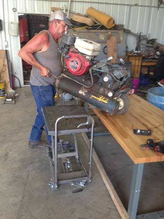 Pictured is an adult male in the process of transferring his air compressor to the worktable from his foot-pumped hydraulic lift cart that’s been raised to the same height.