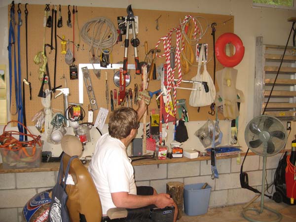 Pictured is an adult male in his power chair observing the many and varied items (e.g., hand tools, cords, totes, measuring instruments) hanging from holders on his large pegboard.