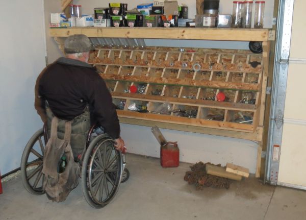 Pictured is an adult male in his manual chair in the process of selecting items in the 60-plus, four-tiered, labeled bins (plus additional items on top) of his homemade storage structure.