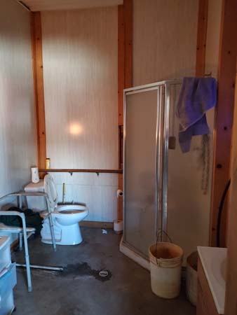 Pictured is a workshop-located restroom that includes enclosed shower, toilet next to a wall with full-width grab-bar, and sink with walker surrounding it to provide stand-up stability.