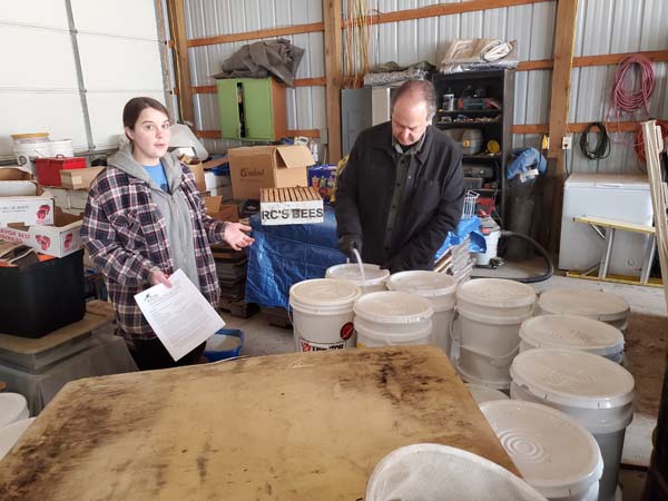 Pictured is a male farmer and female voc-rehab counselor (assessment/plan sheet in hand) in his farm building’s shop-storage area that’s full of various different items.