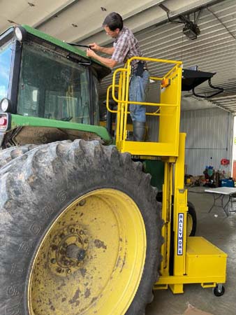 Pictured standing on the guardrail-protected platform of his fully extended moveable lift is an adult male repairing the roof of his tractor’s enclosed cab. 